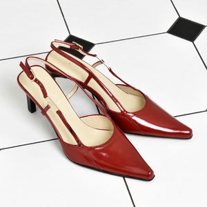 Peut inclure: Une paire d'escarpins slingback en cuir verni rouge foncé avec un bout pointu, une fine bride de cheville réglable avec une boucle dorée et un petit talon noir. La semelle intérieure porte le nom de la marque Gucci en beige clair.