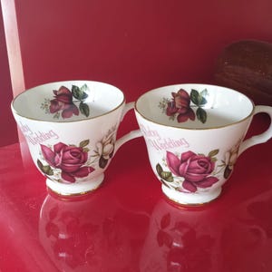 Pode incluir: Duas chávenas de chá em porcelana branca com rebordo e base dourados. Cada chávena está decorada com ilustrações de rosas vermelho-escuras, folhas verdes e o texto Ruby Wedding impresso em vermelho na superfície exterior.