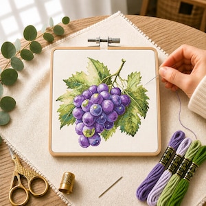 May include: A square wooden embroidery hoop containing a cross-stitch pattern of a cluster of purple grapes with green leaves on white fabric. The set includes four skeins of embroidery floss in purple and green, a gold thimble, and gold scissors.