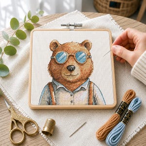 May include: A square wooden embroidery hoop containing a cross-stitch pattern of a brown bear wearing round blue-tinted glasses and a light blue collared shirt with brown suspenders. The kit includes gold scissors, a thimble, and embroidery floss.