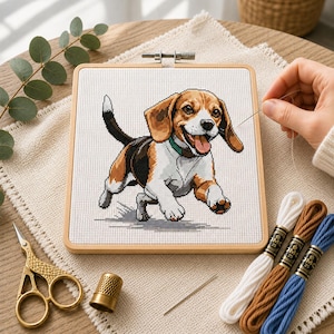 May include: A square wooden embroidery hoop containing white Aida fabric with a cross-stitch pattern of a tricolour beagle dog. The beagle has brown, black, and white fur, a green collar, and is depicted in a running pose with its tongue out.