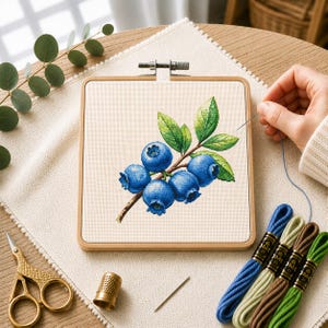 Può includere: Un telaio da ricamo quadrato in legno contenente un tessuto color crema con un disegno a punto croce di un ramo di mirtilli con cinque bacche blu e foglie verdi, accompagnato da fili da ricamo, un ditale e forbici dorate.