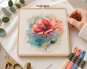 May include: A square wooden embroidery hoop containing a cross-stitch project of a red poppy flower with watercolor-style blue and green splashes. The kit includes gold-toned scissors, a metal thimble, a needle, and four skeins of embroidery floss.