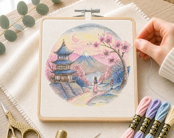 Op de afbeelding: Een vierkant houten borduurraam met een kruissteekontwerp van een blauwe pagode, een berg en roze kersenbloesems. De set bevat een goudkleurige ooievaarsschaar, een vingerhoed, een naald en vier strengen borduurgaren in pastelkleuren.