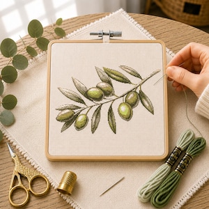 Può includere: Un telaio da ricamo quadrato in legno contenente un motivo a punto croce di un ramo d'ulivo con olive e foglie verdi su tessuto color crema. Sono inclusi forbici dorate, un ditale, un ago da cucito e due matassine di filo da ricamo verde.