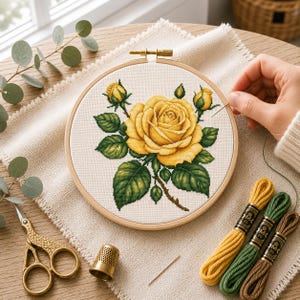 May include: A round wooden embroidery hoop containing a cross-stitch pattern of a large yellow rose with green leaves and two buds. Beside the hoop are three skeins of embroidery floss in yellow, green, and brown, a gold thimble, and gold scissors.