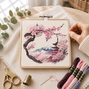 May include: A square wooden embroidery hoop containing a cross-stitch project of a landscape with a mountain, a pagoda, and cherry blossom trees. The design features pink, white, and dark brown threads. Includes embroidery floss, a needle, and scissors.