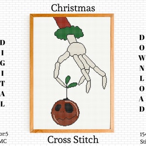 Puede incluir: Patrón de punto de cruz de una mano esquelética sosteniendo un adorno navideño. El adorno es una esfera marrón con un tallo verde y una cara de calabaza. El patrón tiene 154 puntos de ancho por 200 puntos de alto.