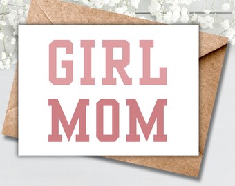 May include: A white card with the text "GIRL MOM" in pink letters. The card is sitting on a brown envelope.