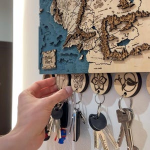 May include: Wooden Middle Earth map key holder with a blue ocean and brown land details. The key holder has several round keychains with different designs. A hand is reaching for one of the keychains.