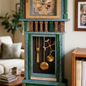 May include: A tall wooden grandfather clock featuring a blue and green painted finish with decorative carved columns. The clock face displays circular text and small portraits, while the lower section reveals gold-toned pendulums and weights.