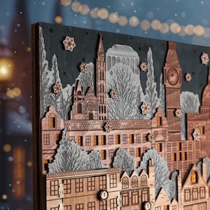 Op de afbeelding: Houten adventskalender met een gedetailleerde stadsgezicht met gebouwen, bomen en sneeuwvlokken. De kalender heeft een donkergrijze achtergrond en is gemaakt van gelaagd hout met ingewikkelde uitsparingen. De gebouwen omvatten een kerk, een klokkentoren en andere architectonische elementen.