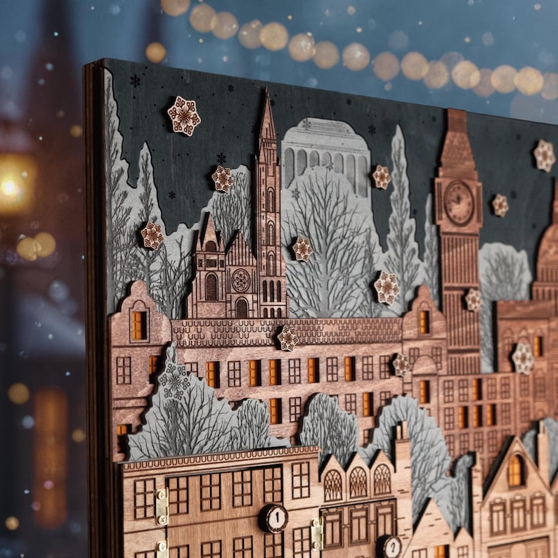 Op de afbeelding: Houten adventskalender met een gedetailleerde stadsgezicht met gebouwen, bomen en sneeuwvlokken. De kalender heeft een donkergrijze achtergrond en is gemaakt van gelaagd hout met ingewikkelde uitsparingen. De gebouwen omvatten een kerk, een klokkentoren en andere architectonische elementen.