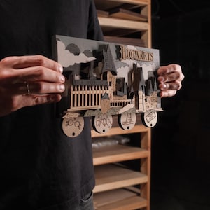 May include: A detailed wooden model of Hogwarts castle, laser-cut with precision. The design includes the word 'Hogwarts', a fence, and castle towers. Circular elements feature glasses, a sorting hat, and a ticket. The color scheme uses grey and natural wood tones.
