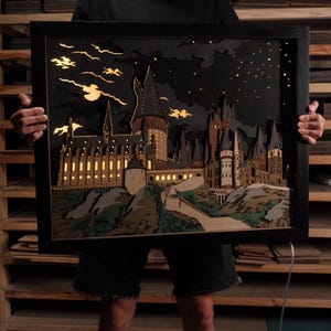 Può includere: Un'opera d'arte in legno a strati incorniciata che raffigura una scena di castello di notte. L'opera presenta un cielo scuro con una luna e stelle, e il castello è illuminato dall'interno. La cornice è nera.