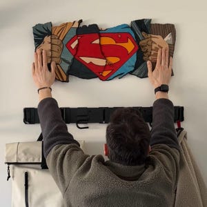 May include: Superman logo wall art with a red and yellow "S" on a blue shield, framed by comic book style hands tearing through the image. A black coat rack is mounted below.