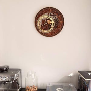 May include: A circular wall clock featuring a dark brown wooden frame with Roman numerals and an intricate, layered gear mechanism. A gold-colored, open-work circular overlay sits off-center on the clock face, with matching gold hands.