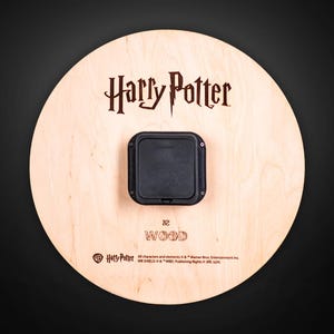 Puede incluir: Un objeto redondo de madera con el tema de Harry Potter con el logotipo de Harry Potter en marr&oacute;n oscuro. Un componente cuadrado negro est&aacute; adherido a la madera. La palabra "WOOD" est&aacute; impresa debajo.