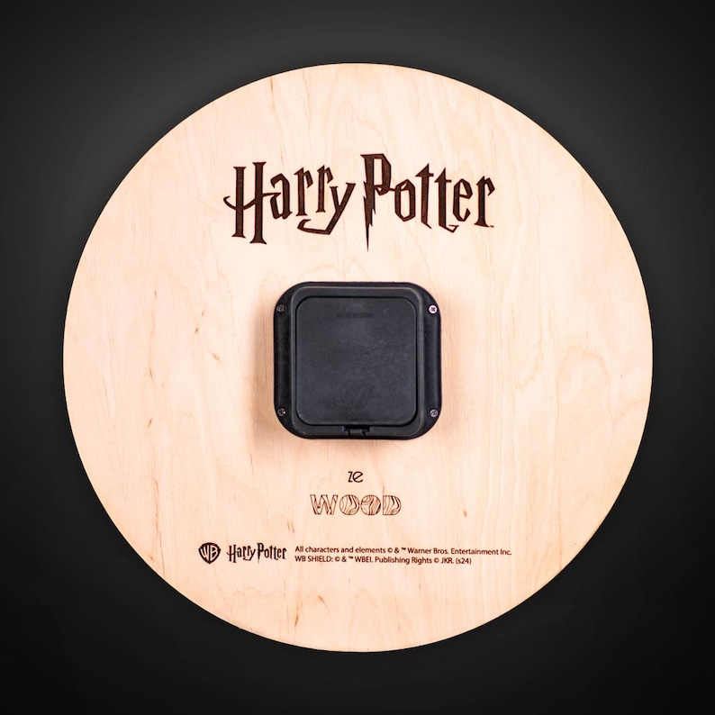 Puede incluir: Un objeto redondo de madera con el tema de Harry Potter con el logotipo de Harry Potter en marr&oacute;n oscuro. Un componente cuadrado negro est&aacute; adherido a la madera. La palabra "WOOD" est&aacute; impresa debajo.