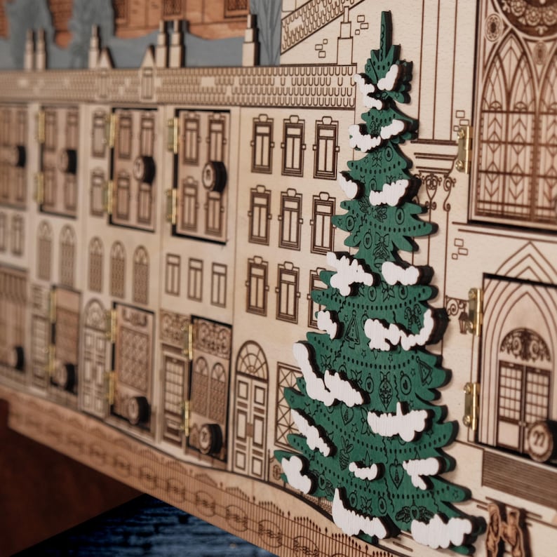 Op de afbeelding: Houten adventskalender in de vorm van een stad, met kleine deuren en een groene kerstboom met witte accenten. De kalender heeft gedetailleerde lasergesneden ontwerpen van gebouwen en architectonische elementen.