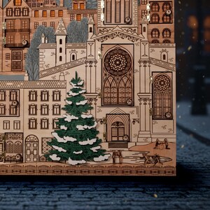 Op de afbeelding: Een houten adventskalender met een gedetailleerd stadsgezichtontwerp. De kalender heeft een groene kerstboom met witte accenten, gebouwen en een kerk. Het ontwerp is lasergesneden.