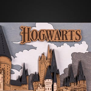 May include: A wooden 3D model of Hogwarts castle from the Harry Potter series. The castle is made of layered wood and features intricate details, including the iconic towers and the Hogwarts crest. The model is painted in shades of brown and gray, and the word "HOGWARTS" is prominently displayed at the top.