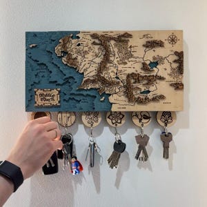 May include: Wooden key holder depicting a detailed map of Middle Earth, with a blue ocean and a detailed landmass. The key holder has several key hooks with decorative wooden keychains. The text "Middle Earth" is visible.