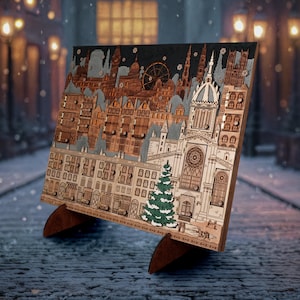 Op de afbeelding: Houten adventskalender met een gedetailleerd stadsgezichtontwerp. Het lasergesneden kunstwerk toont gebouwen, een reuzenrad en een kerk, met een kleine groene kerstboom. De kalender staat op een houten standaard.