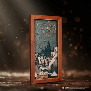 May include: Wooden framed art piece featuring a winter scene with a clock design. The artwork depicts a snowy village with a castle in the background, and the text "HOGSMEADE HOLIDAYS". The frame is a warm brown color.