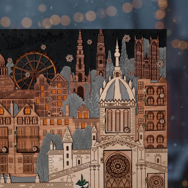 Op de afbeelding: Houten adventskalender met lasergesneden ontwerp van een stadsgezicht. De kalender toont gebouwen, een reuzenrad en een kerk, met genummerde deurtjes voor elke dag. Het kleurenpalet omvat bruin, grijs en cr&egrave;metinten.