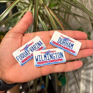 May include: Three white rectangular magnets with blue borders, each featuring a different North Carolina city name: Durham, Fuquay-Varina, and Wilmington. The magnets have a license plate design with the words "First in Flight" at the top.