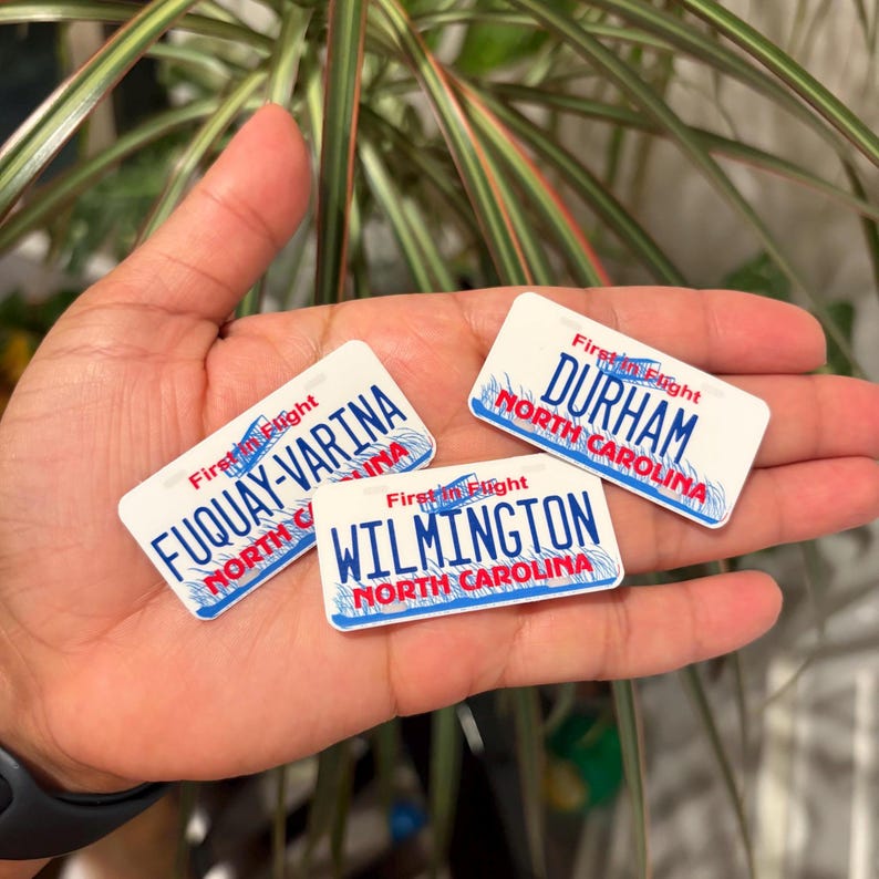 May include: Three white rectangular magnets with blue borders, each featuring a different North Carolina city name: Durham, Fuquay-Varina, and Wilmington. The magnets have a license plate design with the words "First in Flight" at the top.