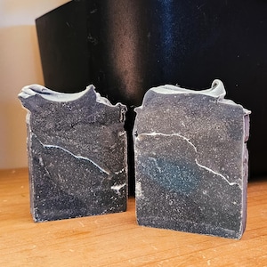 May include: Two dark gray, rectangular soap bars with a textured surface and white veining sit on a wooden surface. A black pot with a small evergreen tree is in the background.