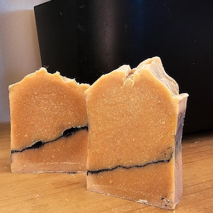 May include: Two handmade soap bars with a light brown color and a dark line running through them. The soaps are on a wooden surface, with a black container and green plant in the background.