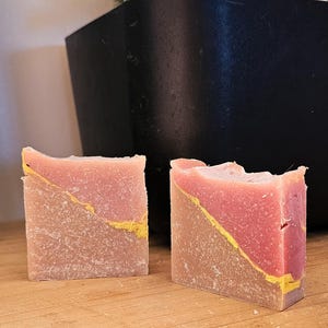 May include: Two handmade soaps with a two-tone design. The top half is a light red colour, and the bottom half is a light brown colour. A yellow line runs diagonally across the soap. The soaps are on a wooden surface.