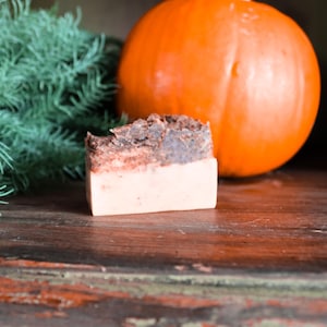 May include: A bar of handmade soap with a brown and beige swirl design. The soap is sitting on a wooden surface next to a large orange pumpkin.