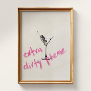 Op de afbeelding: Een ingelijste kunstdruk met een zwart-witillustratie van een martiniglas met drie olijven aan een prikker. Onder het glas staat de tekst "extra dirty please" in een felroze, handgetekend lettertype dat lijkt op een penseelstreek.