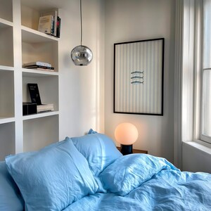 K&ouml;nnte beinhalten: Ein Schlafzimmer mit hellblauer Bettw&auml;sche, bestehend aus zwei Kissen und einer Bettdecke. Ein gerahmter Druck von drei Fischen auf hellblau-wei&szlig; gestreiftem Hintergrund h&auml;ngt an der Wand &uuml;ber einer kugelf&ouml;rmigen Tischlampe mit warmem Licht.