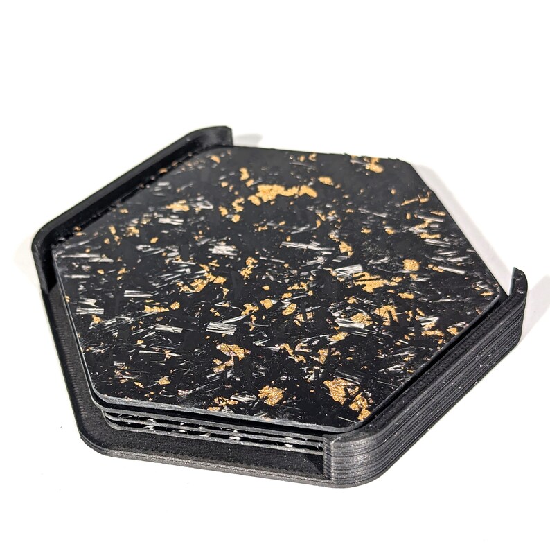 May include: A set of four black hexagonal coasters with a gold flecked pattern. The coasters are stacked on top of each other.