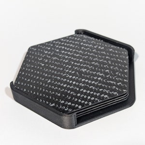 May include: A set of six black hexagonal coasters with a white carbon fiber pattern. The coasters are stacked and held together by a black plastic frame.