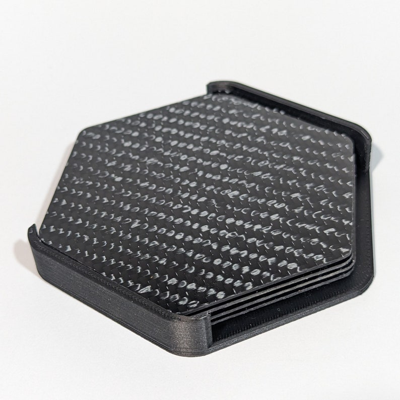 May include: A set of six black hexagonal coasters with a white carbon fiber pattern. The coasters are stacked and held together by a black plastic frame.