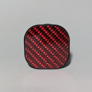 May include: A red and black square-shaped bike reflector with a carbon fiber pattern.