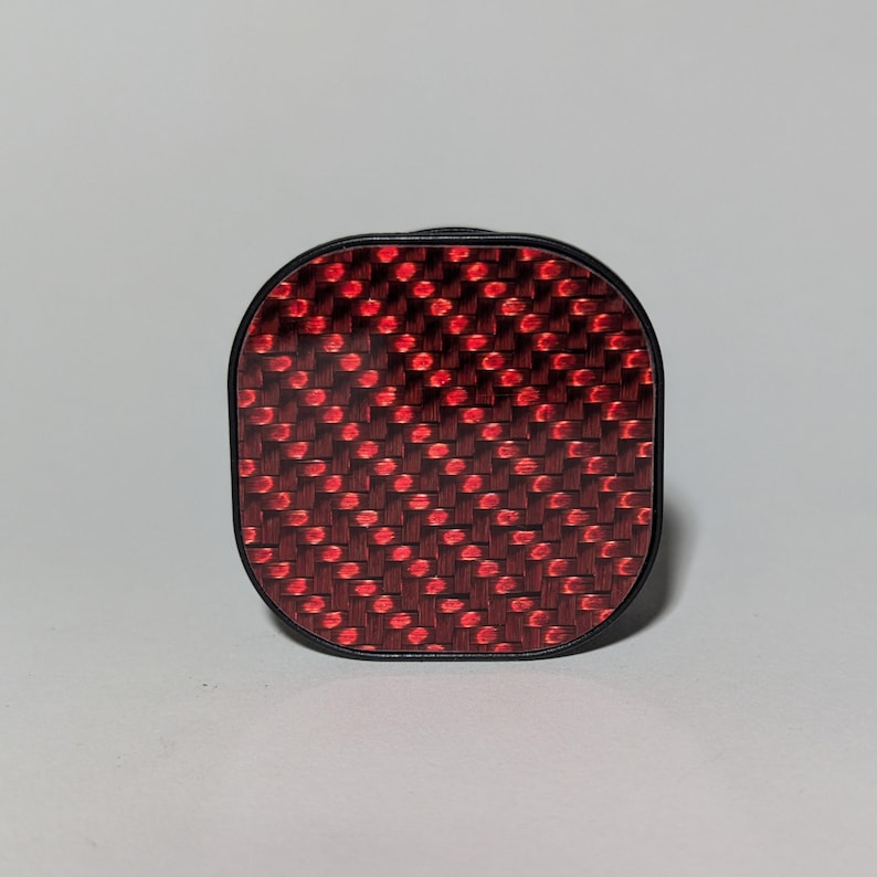 May include: A red and black square-shaped bike reflector with a carbon fiber pattern.