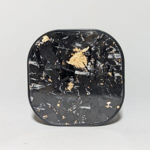 May include: A black square-shaped phone charger with a gold flecked carbon fiber design.
