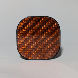 May include: A square-shaped phone grip with a black border and a brown and orange carbon fiber pattern.