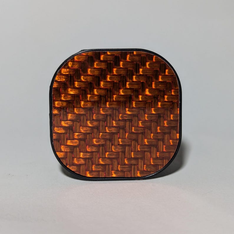 May include: A square-shaped phone grip with a black border and a brown and orange carbon fiber pattern.