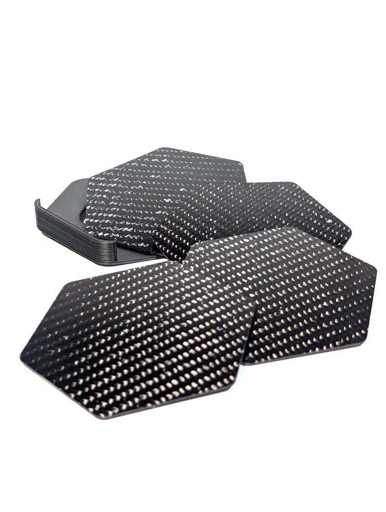 May include: A set of six black hexagonal coasters with a carbon fiber pattern. The coasters are stacked on top of each other.