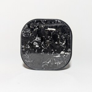 May include: A black square-shaped phone charger with a carbon fiber pattern.