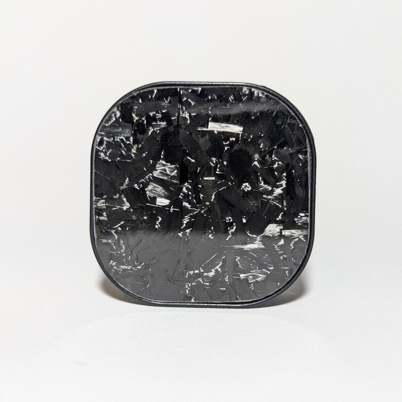 May include: A black square-shaped phone charger with a carbon fiber pattern.