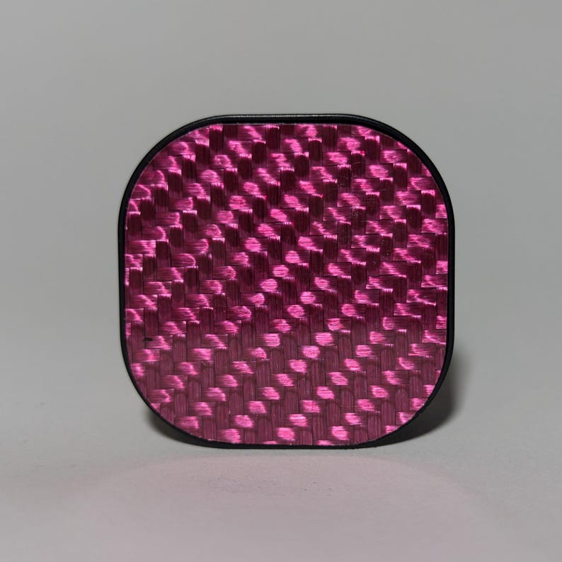 May include: A pink and black square-shaped phone case with a carbon fiber pattern.